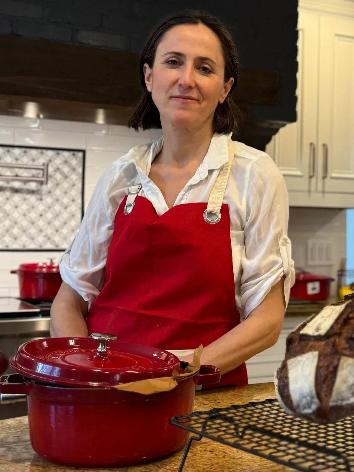 Woman in red apron with freshly baked bread and a red pot in a kitchen.